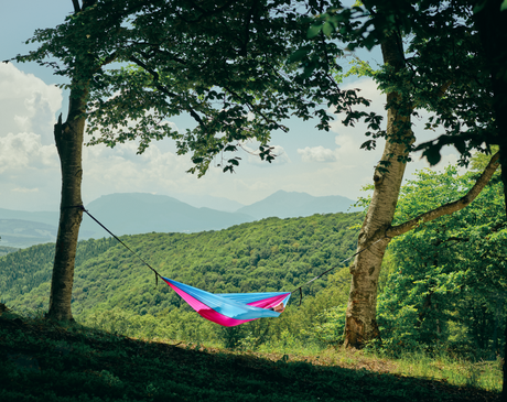 Hamac hikerjoy roz cu bleu montat între doi copaci, peisaj montan