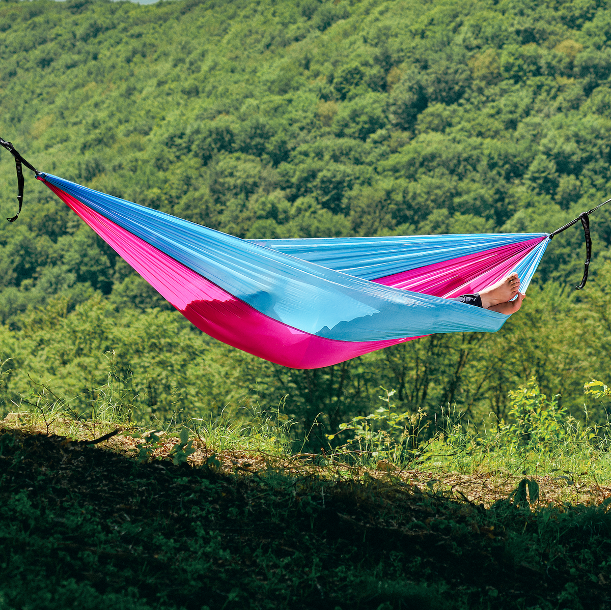 Hamac hikerjoy roz cu bleu montat între copaci, cu pădure în fundal