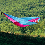 Hamac hikerjoy roz cu bleu montat între copaci, cu pădure în fundal