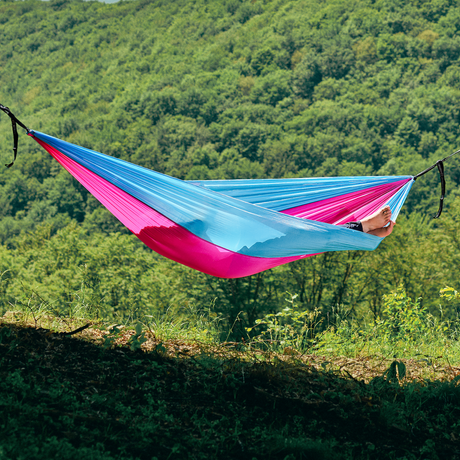 Hamac hikerjoy roz cu bleu montat între copaci, cu pădure în fundal