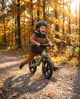 Băiețel cu cască alunecă pe bicicleta fără pedale verde matcha cu roți gonflabile, pe potecă de pădure